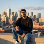 A stylish young man with glasses and a beard sits confidently on a concrete rooftop ledge overlooking a vibrant city skyline at sunset. He is wearing an oversized black knit sweater, wide-leg stonewashed jeans, and chunky white sneakers. A glass of a lime beverage with a straw rests on the ledge next to him.