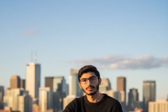 A stylish young man with glasses and a beard sits confidently on a concrete rooftop ledge overlooking a vibrant city skyline at sunset. He is wearing an oversized black knit sweater, wide-leg stonewashed jeans, and chunky white sneakers. A glass of a lime beverage with a straw rests on the ledge next to him.