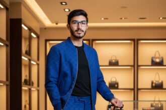 4K ultra-realistic DSLR photo of a handsome Asian man in a royal blue Gucci monogram jacket and pants, black T-shirt, and sneakers, standing next to a luggage cart with a black Gucci backpack and a trolley in a high-end luxury boutique with designer bags displayed.