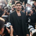 A man with dark hair and glasses, wearing an oversized black blazer over a black tank top, walks through a crowd of paparazzi. Multiple camera flashes are visible, creating a dramatic, celebrity-like atmosphere with daylight shining from the side.