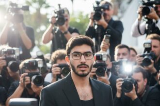 A man with dark hair and glasses, wearing an oversized black blazer over a black tank top, walks through a crowd of paparazzi. Multiple camera flashes are visible, creating a dramatic, celebrity-like atmosphere with daylight shining from the side.