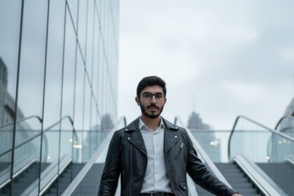 Full-body, cinematic photo of a stylish man in a black biker jacket, white shirt, and dark jeans walking up an outdoor glass-and-metal escalator with a dynamic pose.