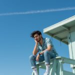 Low-angle vertical portrait of a young man with dark messy hair and glasses, seated pensively on a mint green lifeguard stand against a bright blue sky. He wears a soft blue Hawaiian shirt, cuffed jeans, and white sneakers, conveying retro summer nostalgia.