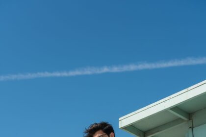 Low-angle vertical portrait of a young man with dark messy hair and glasses, seated pensively on a mint green lifeguard stand against a bright blue sky. He wears a soft blue Hawaiian shirt, cuffed jeans, and white sneakers, conveying retro summer nostalgia.