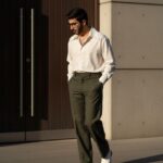Full-body, high-fashion editorial photo of a tall man walking in an urban setting. He wears a creamy white oversized shirt with a wide collar, tucked into high-waisted olive green wide-leg trousers, and white dress boots. The lighting is dramatic, high-contrast late afternoon sun.