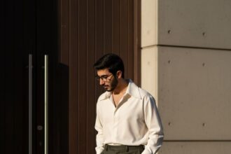 Full-body, high-fashion editorial photo of a tall man walking in an urban setting. He wears a creamy white oversized shirt with a wide collar, tucked into high-waisted olive green wide-leg trousers, and white dress boots. The lighting is dramatic, high-contrast late afternoon sun.