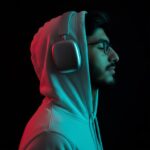 Side profile portrait of person in hood with Apple silver over-ear headphones, lit by cyan neon key light and red rim light.