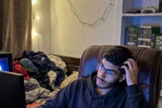 Young man in black hoodie gaming late at night with RGB keyboard, dual monitor setup, and energy drinks on desk
