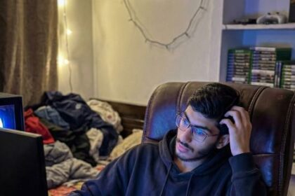 Young man in black hoodie gaming late at night with RGB keyboard, dual monitor setup, and energy drinks on desk
