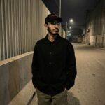 Young man in black jacket and olive cargo pants standing in industrial alley at night with street lights