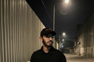 Young man in black jacket and olive cargo pants standing in industrial alley at night with street lights