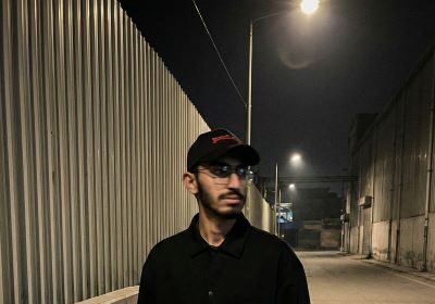 Young man in black jacket and olive cargo pants standing in industrial alley at night with street lights